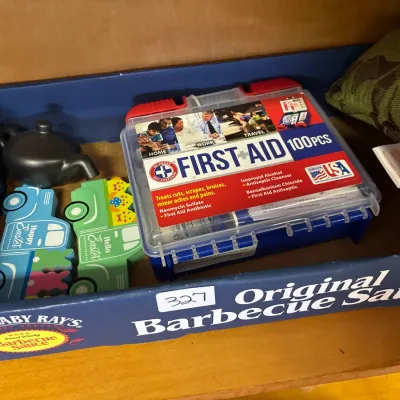 Box of assorted household items including a 100-piece Be Smart Get Prepared first aid kit, decorative wooden Easter trucks, and a small...