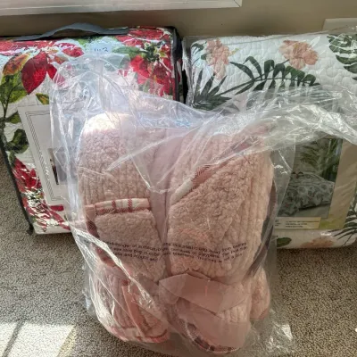 Collection of packaged bedding items including a plush pink fleece throw blanket in plastic wrap alongside two floral quilt sets in...