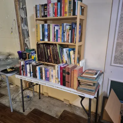Sale Photo Thumbnail #8: Large collection of hardcover and paperback books organized on a light wood bookshelf and a white plastic folding table.