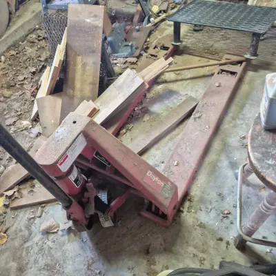 Sale Photo Thumbnail #90: Red Dayton hydraulic pallet jack with visible wear and surface dust, stored in a workshop setting among lumber and tools.