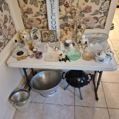 Sale Photo Thumbnail #47: Folding table displaying various home decor items, glass bowls, angel figurines, and kitchenware, with a metal tub and small kettle barbecue underneath.