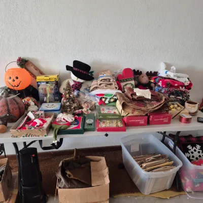 Sale Photo Thumbnail #11: Folding table laden with assorted holiday decor including Halloween jack-o'-lanterns, Christmas snowmen, boxed ornaments, and textiles, alongside storage tubs and boxes below.