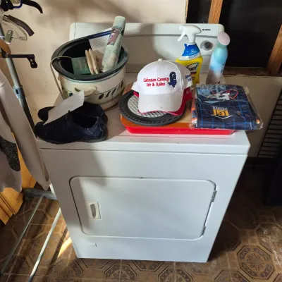 Sale Photo Thumbnail #86: White electric clothes dryer displaying various household items, including a crockpot, cleaning supplies, shoes, and a baseball cap.