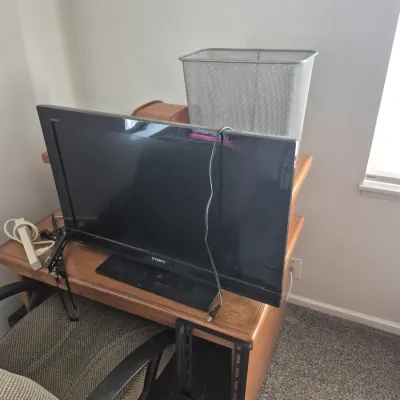 Sale Photo Thumbnail #77: Black Sony Bravia LCD television resting on a wooden desk alongside a silver mesh trash bin and wall mounting bracket.
