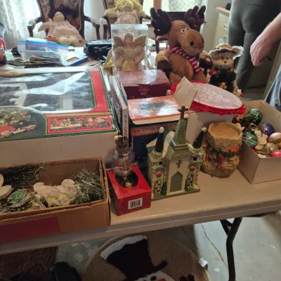 Sale Photo Thumbnail #28: Collection of holiday decor and vintage collectibles displayed on a folding table, including Cabbage Patch Kids dolls, Christmas train set in box, plush moose, and village church figurine.