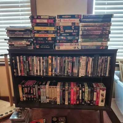 Sale Photo Thumbnail #32: Large collection of VHS tapes and DVDs neatly organized on and inside a dark wood mid-century style bookcase.