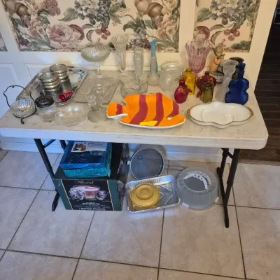 Sale Photo Thumbnail #50: Folding table displaying assorted glassware, decorative ceramics including a bright fish platter, colorful bottles, and kitchen accessories.