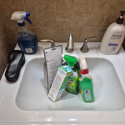 Sale Photo Thumbnail #83: Assorted household supplies including spray cleaners, Aveeno body wash, a Fleet enema box, and a Glacier Bay shower hose resting around a bathroom sink.