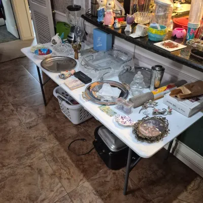 Sale Photo Thumbnail #44: Folding table displaying assorted household items, including silver-tone serving trays, vintage Easter rabbit figurines, glassware, and small decorative home goods.