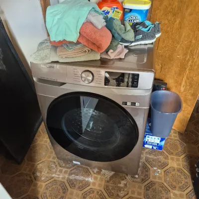 Sale Photo Thumbnail #85: Modern Samsung front-load washing machine in a champagne finish, topped with folded towels, athletic shoes, and laundry supplies.