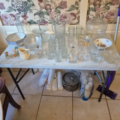 Sale Photo Thumbnail #49: Assorted glassware, serveware, and kitchen items arranged on a folding table, including glass pitchers, drinking glasses, porcelain bowls, and small figurines.