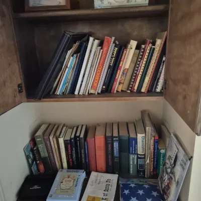Sale Photo Thumbnail #76: Collection of books and cookbooks stored in a wooden cabinet, including textbooks, recipe books, framed decor, and miscellaneous items.