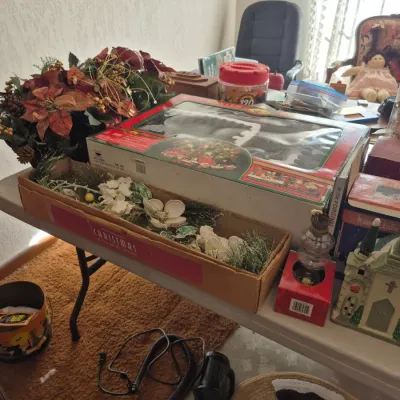 Sale Photo Thumbnail #13: Collection of Christmas decor items on a folding table, including boxed train set, faux floral arrangements, and holiday village pieces.
