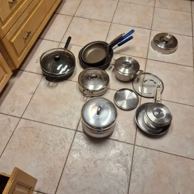 Sale Photo Thumbnail #52: Assorted collection of cookware, including stainless steel stock pots with lids, nonstick frying pans, and glass lids, laid out on a tile floor.