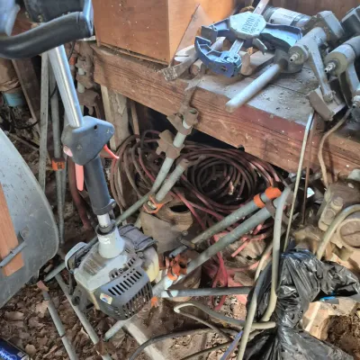 Sale Photo Thumbnail #93: Gas-powered string trimmer resting against a cluttered workshop bench alongside pipe clamps, power tools, and air hoses.