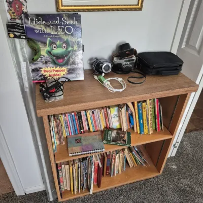 Sale Photo Thumbnail #73: Light oak finish two-shelf bookcase filled with a collection of children's books and vintage paperbacks. The top surface displays electronics including a 35mm camera, camcorder, and a children's puppet book.