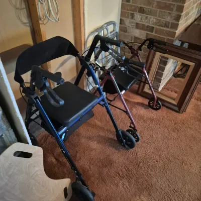 Sale Photo Thumbnail #26: Selection of mobility aids, including two four-wheeled rollator walkers with seats and hand brakes, a standard aluminum walker, and a wooden transfer board.