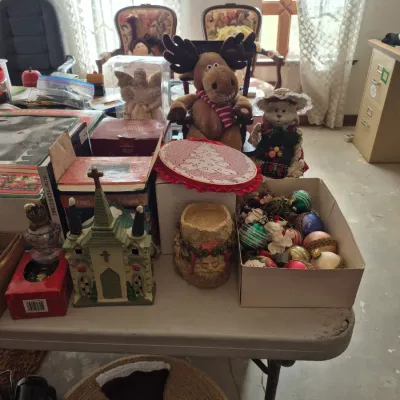 Sale Photo Thumbnail #12: Collection of Christmas decor items on a table, including a plush moose, a miniature church structure, boxed ornaments, and festive figurines.