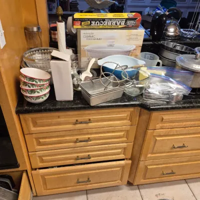 Sale Photo Thumbnail #53: Assorted kitchenware and cookware arranged on a dark granite countertop above light wood cabinets, including Pyrex mixing bowls, loaf pans, a ceramic food warmer in its original box, and Christmas-patterned bowls.