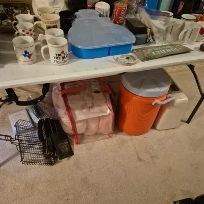 Sale Photo Thumbnail #71: Assorted household goods and kitchenware displayed on and under a folding table, including coffee mugs, a food processor, a water cooler jug, and a snowman-shaped storage tray.