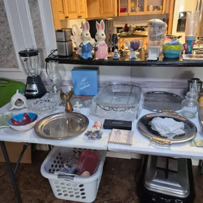 Sale Photo Thumbnail #10: Collection of household goods, including ceramic Easter bunny statues, an Osterizer blender, silver-tone serving trays, glassware, and small kitchen items.
