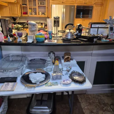 Sale Photo Thumbnail #9: Assorted kitchenware and home decor items displayed on a white folding table and kitchen counter, including serving trays, small knick-knacks, a tea kettle, and countertop appliances.