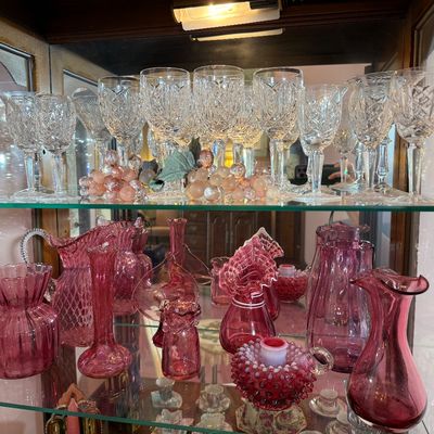 Display cabinet filled with vintage glassware, including cut crystal wine stems on the top shelf and various pink cranberry glass vases,...