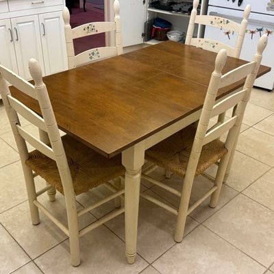Country cottage style dining set featuring a medium wood-tone extendable table and four off-white ladder-back chairs with rush seats.