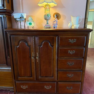Traditional dark wood armoire chest with raised panel cabinet doors and batwing brass bail handles, decorated on top with a porcelain...