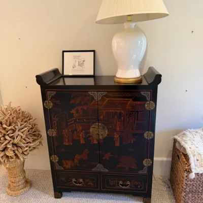 Black lacquer Asian-style cabinet featuring hand-painted figures and landscapes with decorative brass hardware, topped with a white...