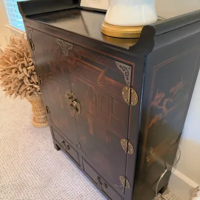 Asian-style black lacquer cabinet featuring hand-painted Chinoiserie landscape scenes, decorative brass hardware, and a raised upturned...