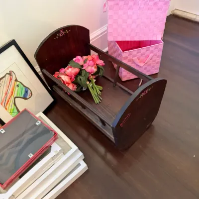 Vintage dark wood doll cradle with painted floral details, accompanied by faux pink flowers, bright pink woven storage bins, and assorted...