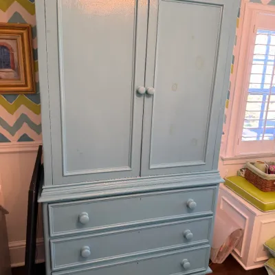 Light blue painted armoire chest with double cabinet doors over three drawers, featuring wooden knobs and bun feet.