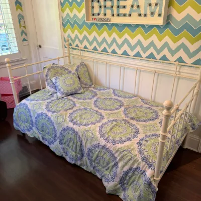 White metal daybed with spindle rails, dressed in blue and green medallion pattern bedding, accompanied by a illuminated wall sign...