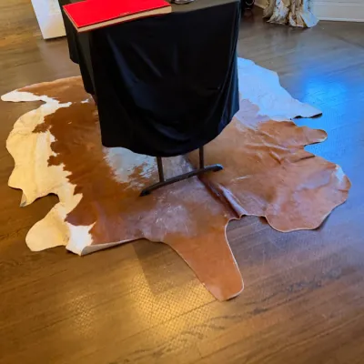 Sale Photo Thumbnail #170: Large natural brown and white cowhide area rug laid out on a hardwood floor beneath a draped display table.