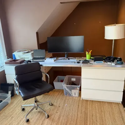 Modern home office setup featuring a white modular desk, black faux leather rolling chair, wide-screen computer monitor, and various desk...