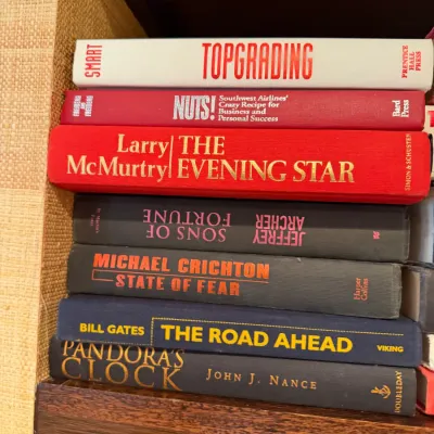 Sale Photo Thumbnail #252: Stack of modern hardcover books on a wooden shelf, including titles by Larry McMurtry, Michael Crichton, and Bill Gates.