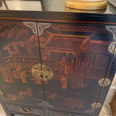 Black lacquered Asian-style cabinet featuring hand-painted red and gold figurative scenes, decorative brass corner brackets, and a...