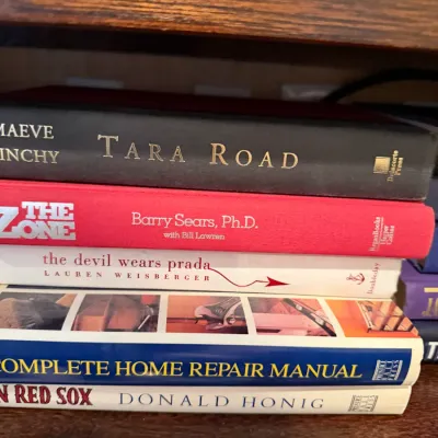 Sale Photo Thumbnail #256: Stack of hardback books including modern fiction, health, and home repair titles, displayed on a wooden shelf.