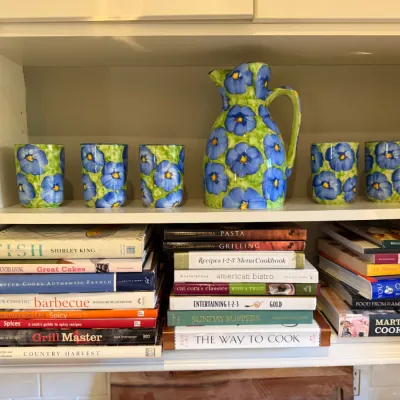 Sale Photo Thumbnail #183: Hand-painted floral pitcher and six matching cups displayed above stacked cookbooks on white open shelving.