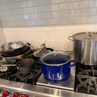 Sale Photo Thumbnail #178: Collection of kitchen cookware on a stove, featuring a blue enameled stock pot, a large stainless steel stock pot, and various frying pans, strainers, and saucepans.