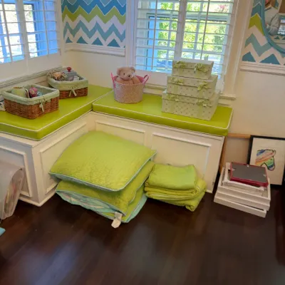 Corner window bench with green cushions, stack of green quilts, woven storage baskets, decorative boxes, and picture frames.