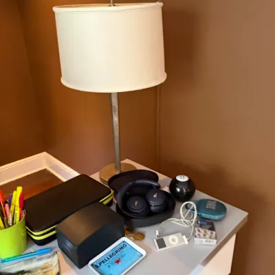 Sale Photo Thumbnail #104: A collection of personal items and electronics displayed on a light gray nightstand beside a tall brushed metal table lamp with a cream drum shade.