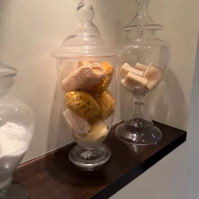 Collection of glass apothecary and apothecary-style jars filled with bath sponges, bar soaps, and salts.