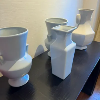 Sale Photo Thumbnail #126: Collection of four modern white ceramic vases in varied geometric shapes arranged on a dark wood table.