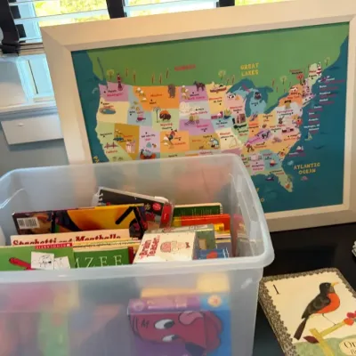 Framed colorful United States illustrated map art alongside a clear plastic storage bin filled with children's books and games.