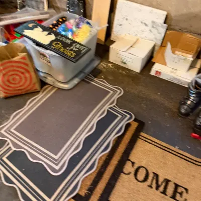 Sale Photo Thumbnail #231: Collection of doormats including scalloped edge mats and a coir 'Welcome' mat, alongside storage bins with seasonal decorations and miscellaneous household items.