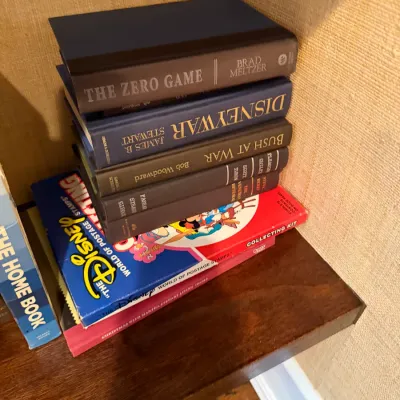 Sale Photo Thumbnail #259: Stack of hardcover books and Disney stamp collecting kits on a wooden shelf.