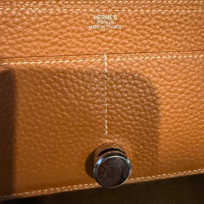 Close-up of a tan Hermès pebbled leather wallet featuring silver-foil branding and an engraved Palladium hardware closure.