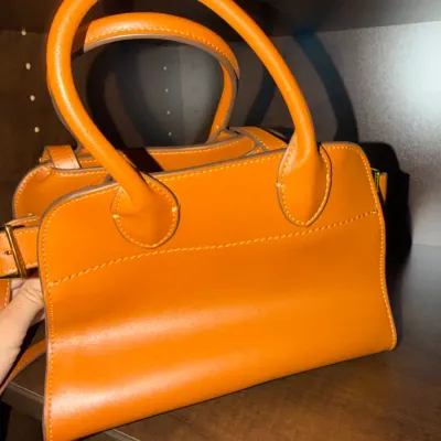 Bright orange structured leather handbag featuring double rounded top handles, tonal stitching, and gold-tone hardware.